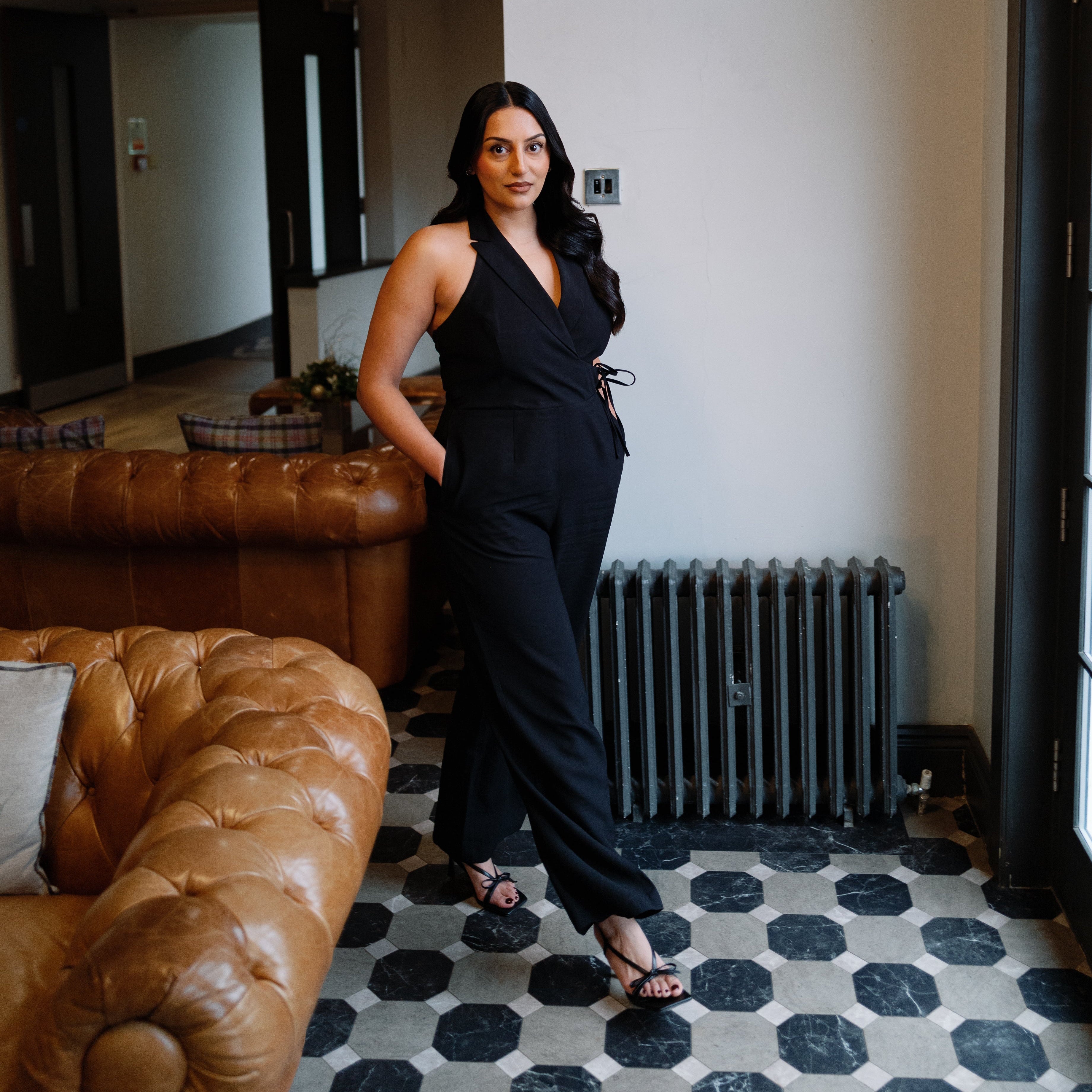 Woman in a black jumpsuit standing in a room with a brown leather couch and hexagonal patterned floor.